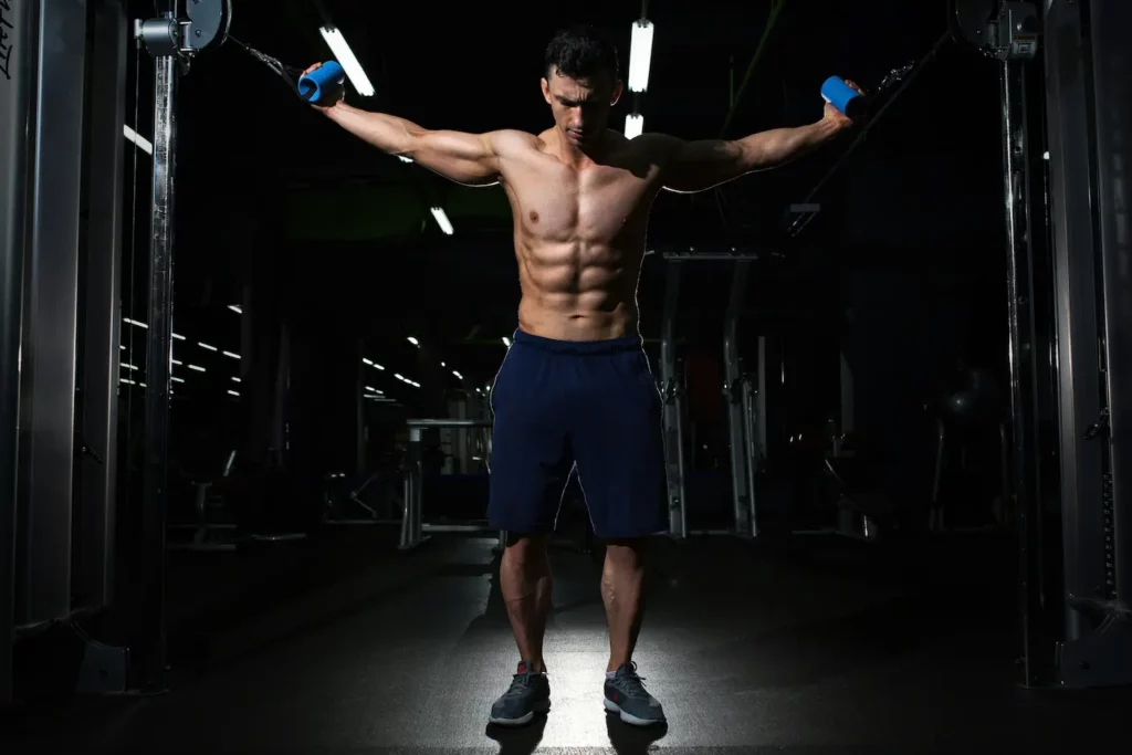 A shirtless man in gym shorts performs a cable fly exercise in a dimly lit gym, showcasing his defined muscles, reminiscent of peak bodybuilding form.